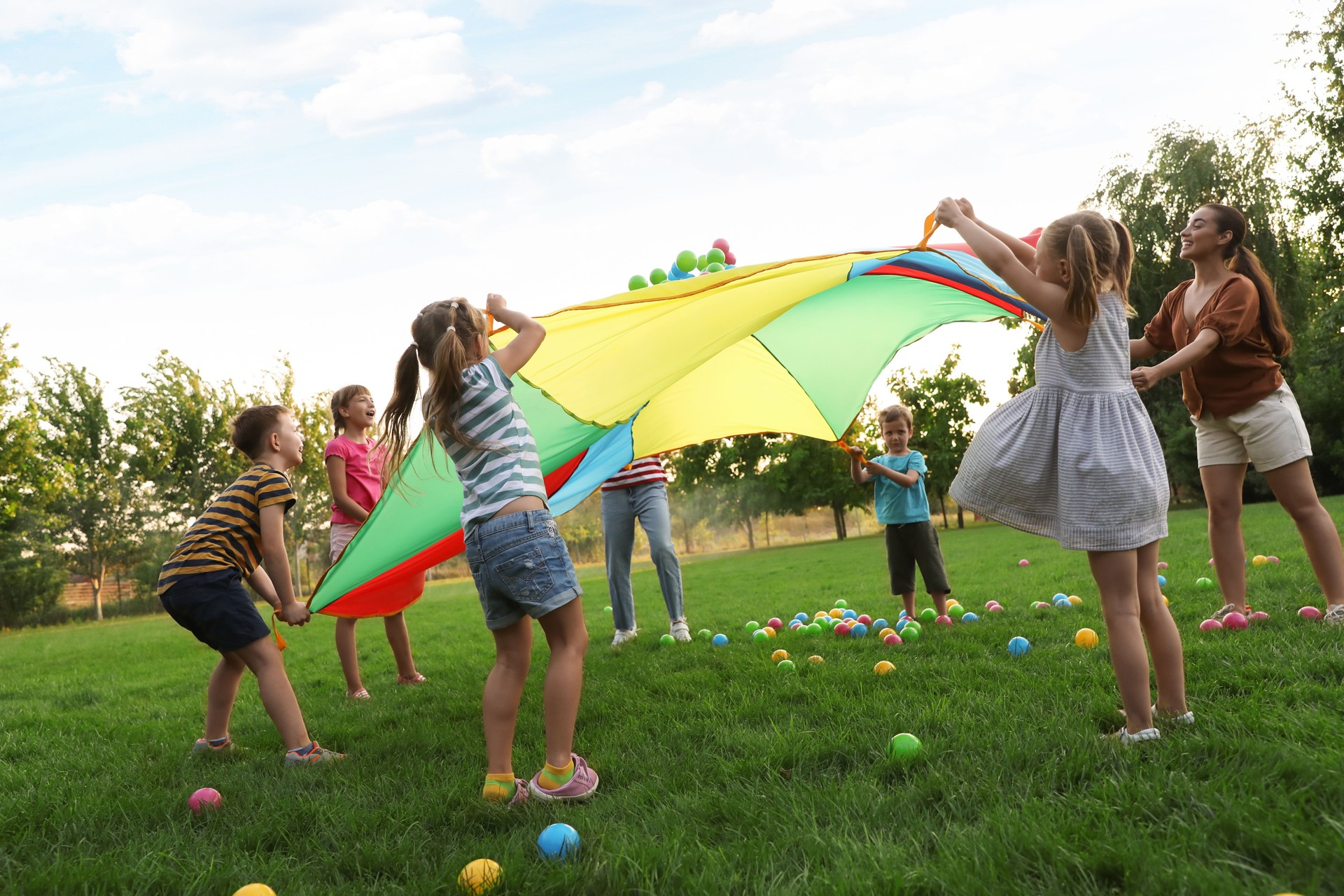 Grupo de niños y profesor jugando con el paracaídas arco iris del parque infantil sobre hierba verde. Actividad de campamento de verano